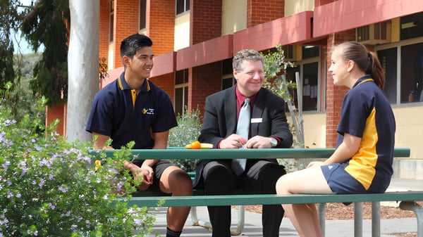 Zwei Schülerinnen und Schüler sitzen mit einer erwachsenen Person auf einer Bank vor einem Backsteingebäude der Paralowie R-12 School.