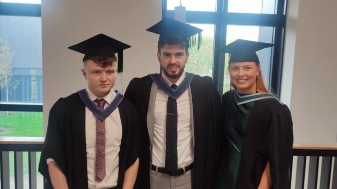 Drei Schüler der Patrician Presentation Secondary School stehen in Abschlusskleidung in einem universitär wirkenden Raum.