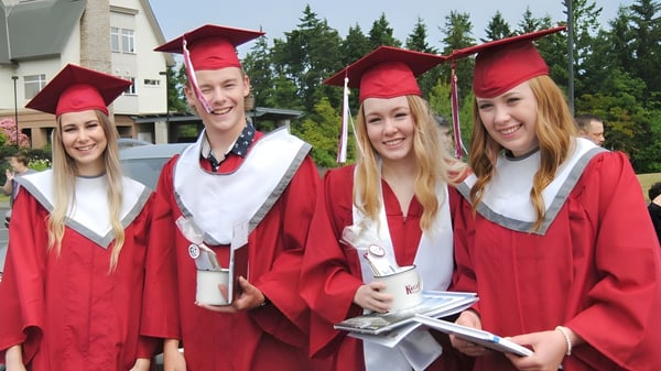 Eine Gruppe von Absolventinnen und Absolventen der Pender Harbour Secondary steht draußen in roten Talaren und Hüten vor einem bewaldeten Hintergrund.
