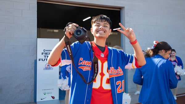 Ein junger Mann in blauem Sporttrikot hält eine Kamera und zeigt ein Peace-Zeichen vor einem Gebäude des Phoenix Union High School District.