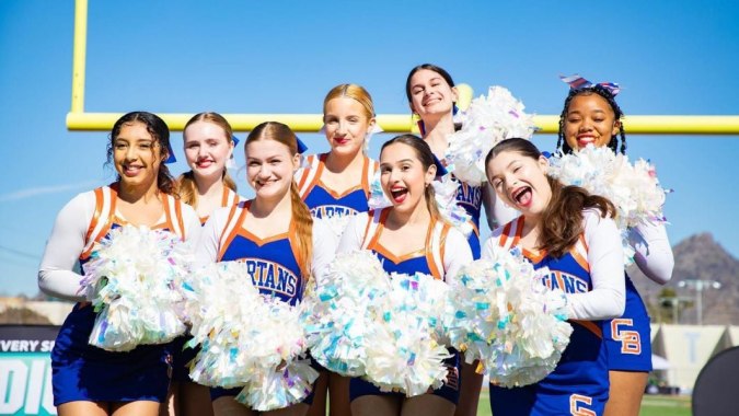 Eine Gruppe Cheerleader der Phoenix Union High School District steht mit bunten Pompons unter blauem Himmel vor einem gelben Torpfosten.