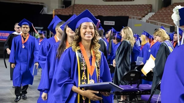 Eine Gruppe von Absolventen der Phoenix Union High School District steht bei einer Indoor-Abschlussfeier in blauen Talaren mit Diplomen.