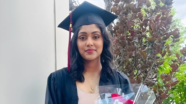 Eine junge Frau in Abschlusskleidung steht vor grünem Hintergrund auf dem Campus der Pine Ridge Secondary School.