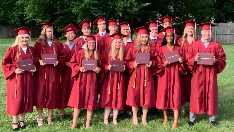 Eine Gruppe von Absolventinnen und Absolventen der Plaza Heights Christian Academy steht im Freien vor grünen Bäumen in roten Abschlussroben.