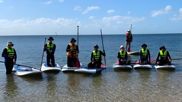 Schüler des Plympton International College stehen mit Rettungswesten auf Paddleboards im flachen Wasser vor ruhigem Ozean.