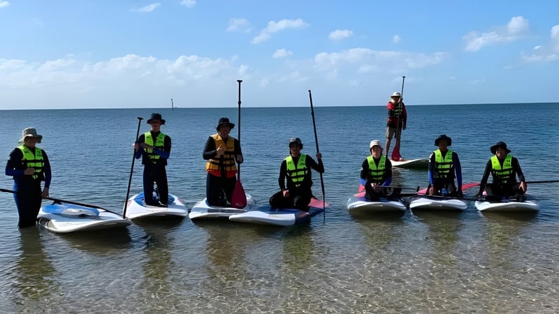 Schüler des Plympton International College stehen mit Rettungswesten auf Paddleboards im flachen Wasser vor ruhigem Ozean.