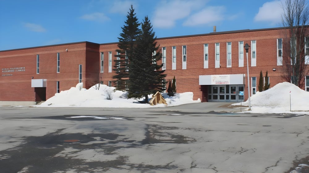 Das große Backsteingebäude der Polyvalente Alexandre-J.-Savoie steht hinter einem hohen Kiefernbaum im schneebedeckten Winter.