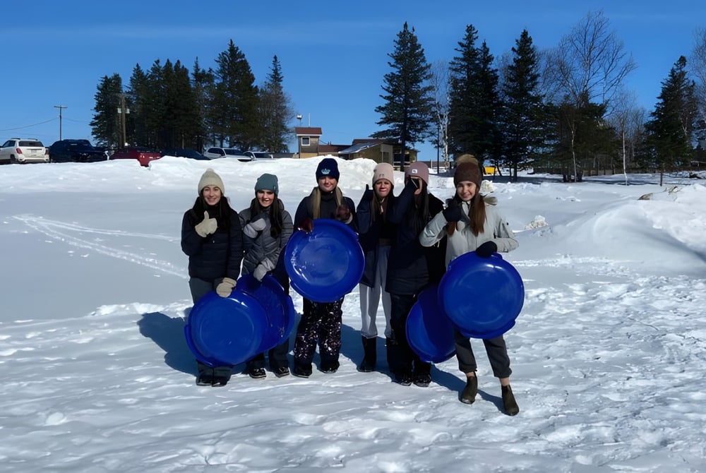 Eine Gruppe Schüler auf einem verschneiten Feld mit Tannenbäumen und Gebäuden im Hintergrund auf dem Campus der Polyvalente Louis-Mailloux.
