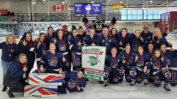 Die Damen-Eishockeymannschaft der Polyvalente Thomas-Albert steht in ihren blauen Trikots in der Eishalle vor kanadischer und britischer Flagge.