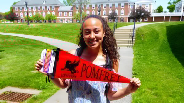 Eine lächelnde junge Frau hält im Gras vor einem Backsteingebäude auf dem Campus der Pomfret School eine rot-weiße Pomfret Fahne.