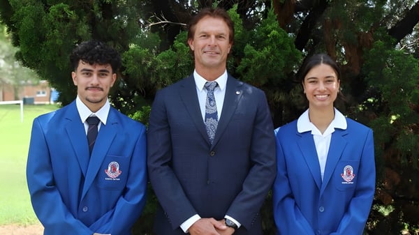 Drei Personen, darunter zwei Schüler in blauen Uniformen, stehen vor einer grünen Kulisse auf dem Gelände der Port Hacking High School.