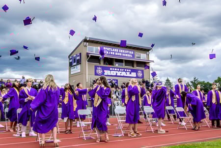 Absolventen der Portland Christian High School feiern mit lila Talaren und fliegenden Hüten auf dem Sportplatz.