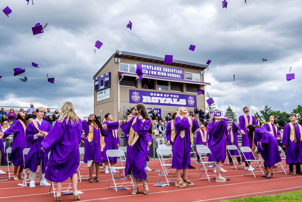 Absolventen der Portland Christian High School feiern mit lila Talaren und fliegenden Hüten auf dem Sportplatz.