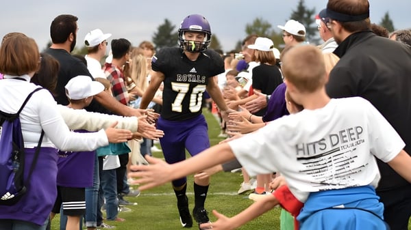 Schüler der Portland Christian High School versammeln sich in Sportuniformen auf einem Spielfeld während Zuschauer im Hintergrund stehen.