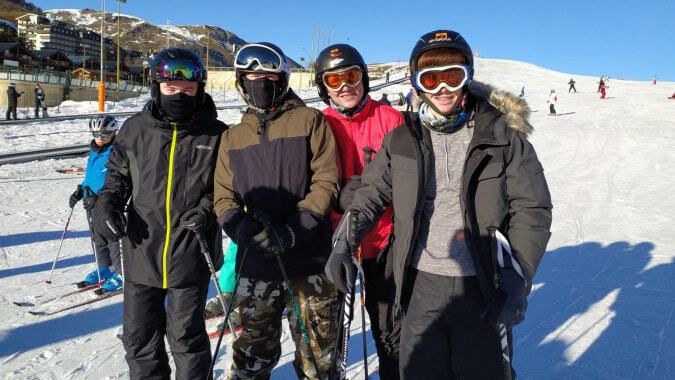 Vier Schülerinnen und Schüler der Portmarnock Community School stehen in Winterkleidung auf einer schneebedeckten Piste mit einem Skigebiet im Hintergrund.
