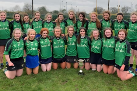 Eine Gruppe weiblicher Fußballspielerinnen vom Portumna Community College posiert auf einem Spielfeld mit einer Trophäe im Vordergrund.