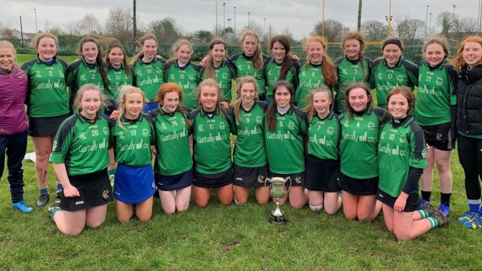 Eine Gruppe junger Fußballspielerinnen in grünen Trikots posiert mit einem Pokal auf dem Spielfeld des Portumna Community College.
