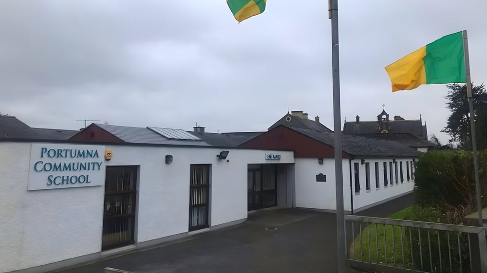 Das Schulgebäude des Portumna Community College mit weißen Außenwänden und grünen sowie gelben Fahnen vor ländlicher Kulisse.