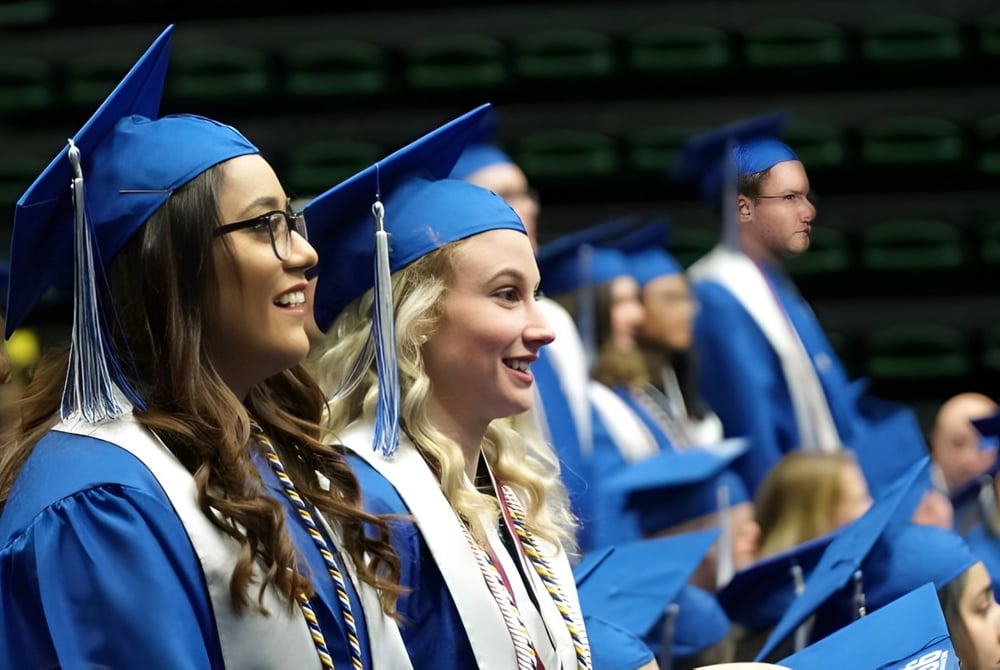 Eine Gruppe von Absolventen in blauen Talaren und Hüten steht gemeinsam vor dunklem Hintergrund auf dem Abschlussfoto des Poudre School District.