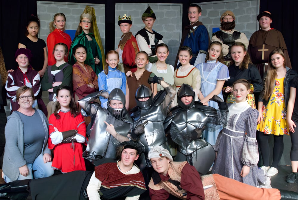 Eine Gruppe Schüler der Prairie Christian Academy posiert zusammen in verschiedenen Kostümen vor einem dunklen Hintergrund mit architektonischen Elementen.