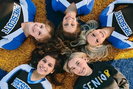 Junge Frauen in Cheerleading-Uniformen liegen lächelnd auf dem Boden vor farbigem Hintergrund auf dem Campus der Prescott High School.