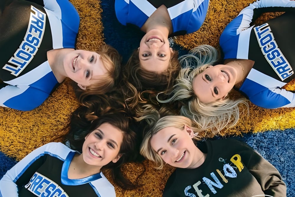 Junge Frauen in Cheerleading-Uniformen liegen lächelnd auf dem Boden vor farbigem Hintergrund auf dem Campus der Prescott High School.
