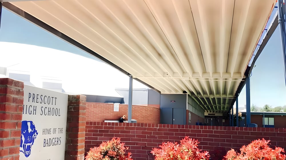 Das Backsteingebäude der Prescott High School mit einem überdachten Weg und orangefarbenen Blumen im Vordergrund unter blauem Himmel.