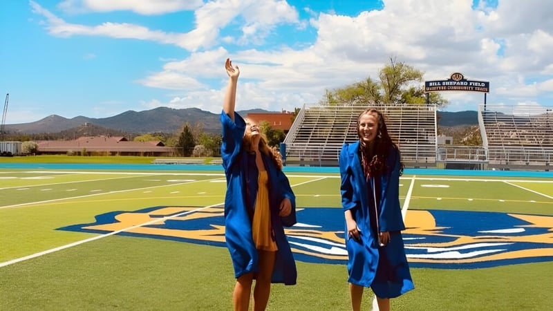 Zwei Personen in blauen Abschlussroben stehen auf dem Sportplatz im Prescott Unified School District vor Bergen und bewölktem Himmel.