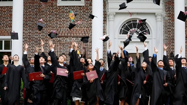 Absolventinnen und Absolventen der Princeton International School of Mathematics & Science werfen ihre Abschlussmützen vor einem Backsteingebäude in die Luft.