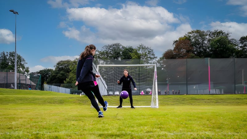 Zwei Frauen in Sportkleidung stehen auf dem Sportplatz von Prior's Field nahe dem Fußballtor unter blauem Himmel.