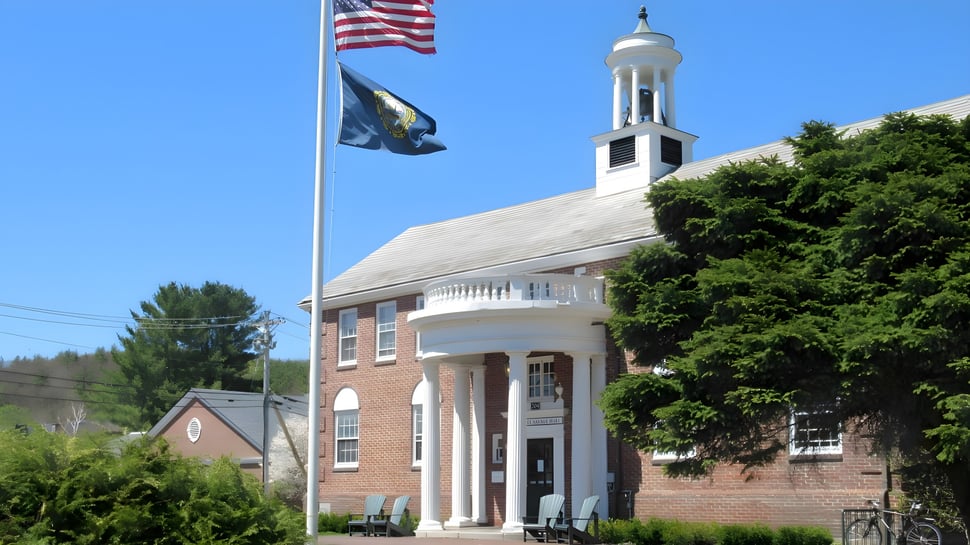 Das Backsteingebäude der Proctor Academy mit einem weißen Turm und zwei wehenden Flaggen ist von Bäumen umgeben.