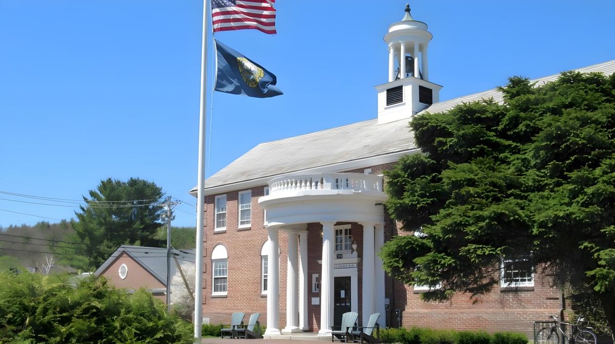 Das Backsteingebäude der Proctor Academy mit einem weißen Turm und zwei wehenden Flaggen ist von Bäumen umgeben.