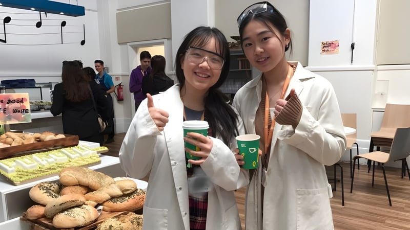 Zwei Schülerinnen der The Purcell School for Young Musicians tragen Laborkittel und halten Kaffeetassen in einem Raum mit Gebäck.