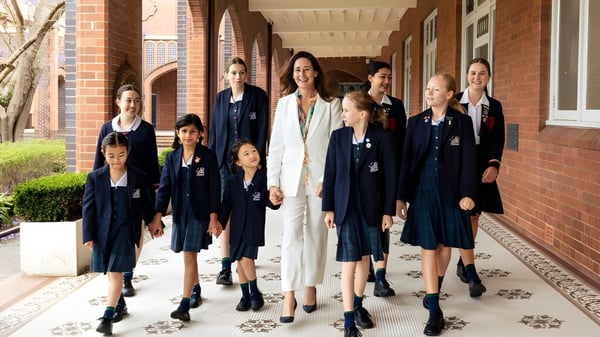 Eine Gruppe von Schülerinnen steht im Flur des Pymble Ladies College vor backsteinernen Wänden und bogenförmigen Fenstern.