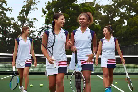 Vier Schülerinnen in Sportkleidung stehen auf dem Tennisfeld des Pymble Ladies College, umgeben von Bäumen.