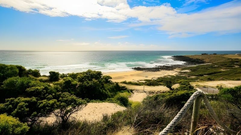 Eine Küstenlandschaft mit Strand, Felsen und Vegetation vor dem ruhigen Meer, aufgenommen in der Nähe von Queechy High School.