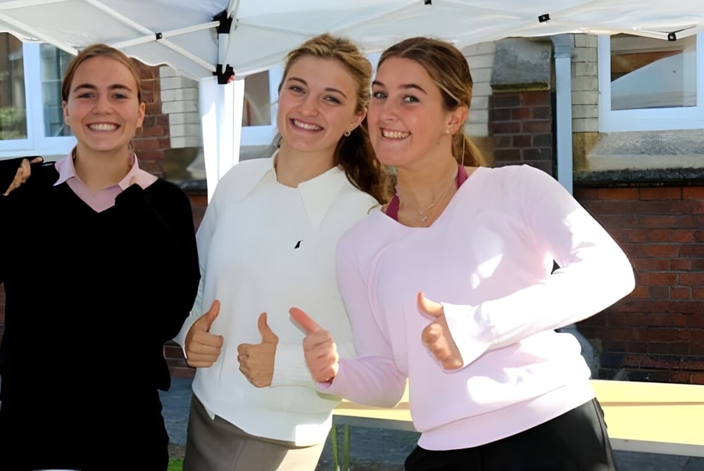 Drei lächelnde Schülerinnen stehen vor einem Gebäude auf dem Campus der Queen Anne’s School.