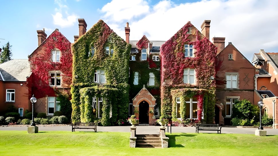 Ein historisches Backsteingebäude mit verzierter Architektur ist von rotem und grünem Laub der Queen Anne’s School umgeben.