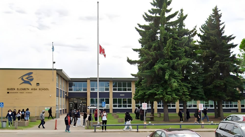 Das Schulgebäude der Queen Elizabeth High School umgeben von hohen Tannen mit einer Gruppe Schülern davor und einem Fahnenmast.