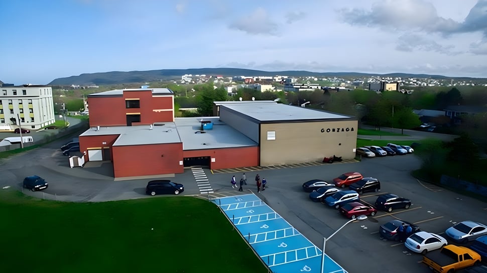 Gebäude der Queen Elizabeth Regional High mit Parkplatz im Vordergrund und Wohngebiet mit Bergen im Hintergrund.