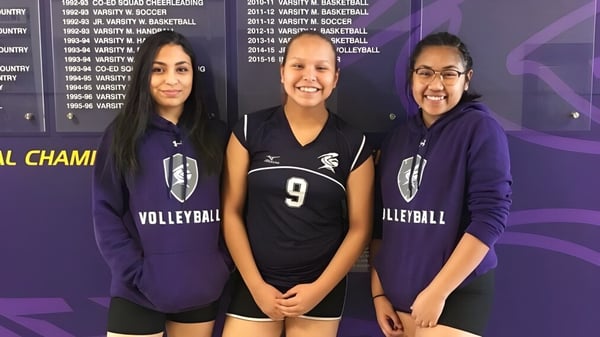 Drei Schülerinnen der Queen Elizabeth High School stehen in ihren Volleyball-Uniformen vor einer Wand mit sportlichen Auszeichnungen.
