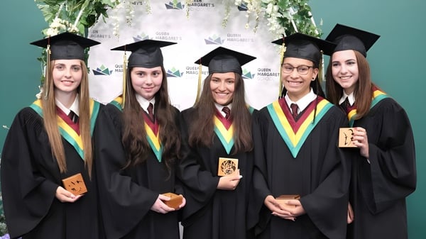 Fünf Schüler der Queen Margaret’s School stehen in ihren Abschlusskutten und halten ihre Diplome vor einem Universitätslogo.