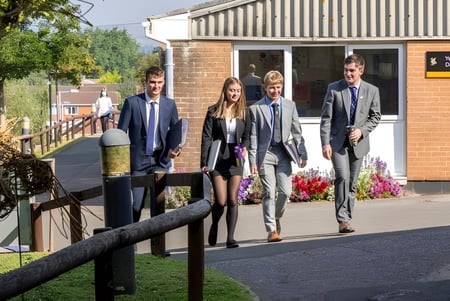 Eine Gruppe von Personen geht entlang eines Weges auf dem Campus des Queen’s College mit begrüntem Umfeld und Gebäuden im Hintergrund.