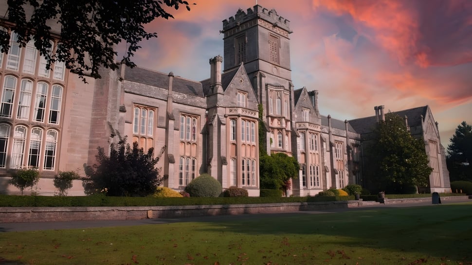 Das prächtige steinerne Hauptgebäude von Queen’s College ist von grüner Natur umgeben und steht vor einem bunten Sonnenuntergangshimmel.