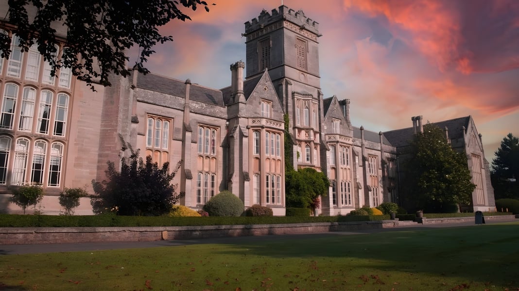 Das prächtige steinerne Hauptgebäude von Queen’s College ist von grüner Natur umgeben und steht vor einem bunten Sonnenuntergangshimmel.