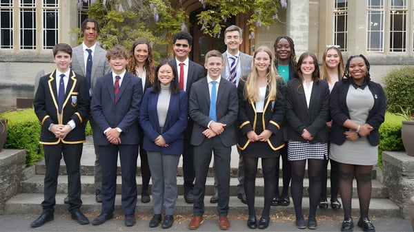 Eine Gruppe von Schülern des Queen’s College posiert in formeller Kleidung auf einer Treppe vor Bäumen.