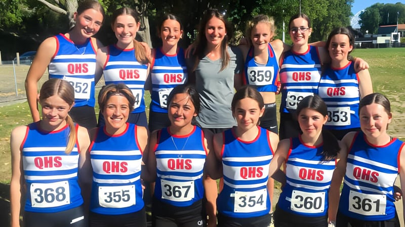 Eine Gruppe Schülerinnen der Queens High School steht in blauen und weißen Sportuniformen auf einem Sportplatz.
