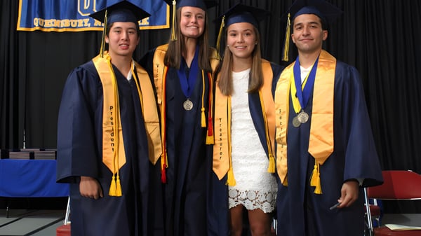 Vier Schüler der Queensbury High School stehen zusammen in Abschlussroben auf einer Bühne mit blau-gelbem Banner im Hintergrund.