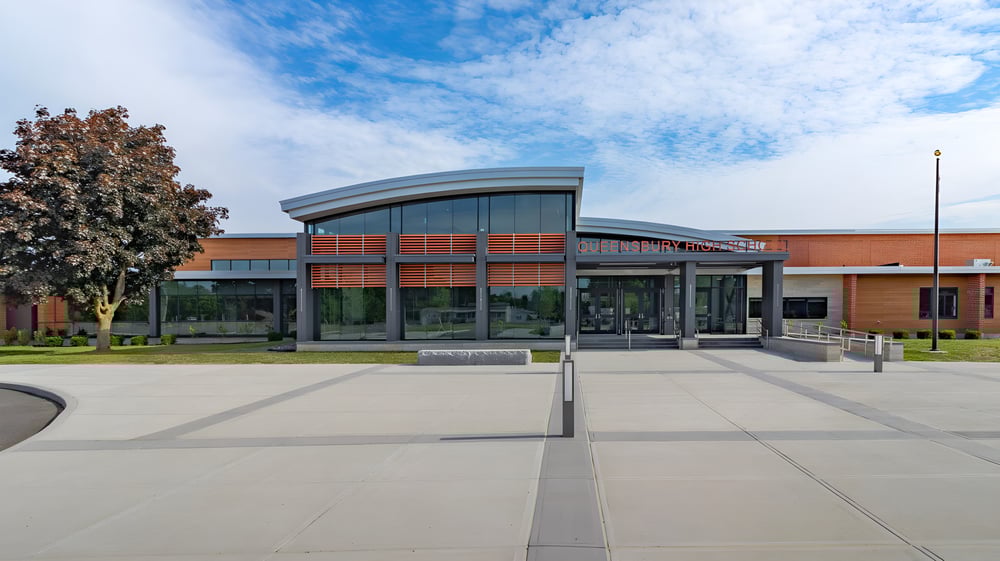 Das moderne Gebäude mit Glasfassade der Queensbury High School steht in einem gepflasterten Innenhof umgeben von Bäumen.