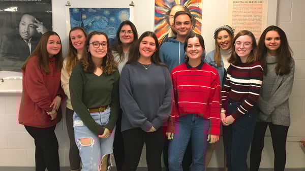 Eine Gruppe Schülerinnen und Schüler steht im Klassenraum des Queensbury Union Free School District vor bunten Postern an der Wand.